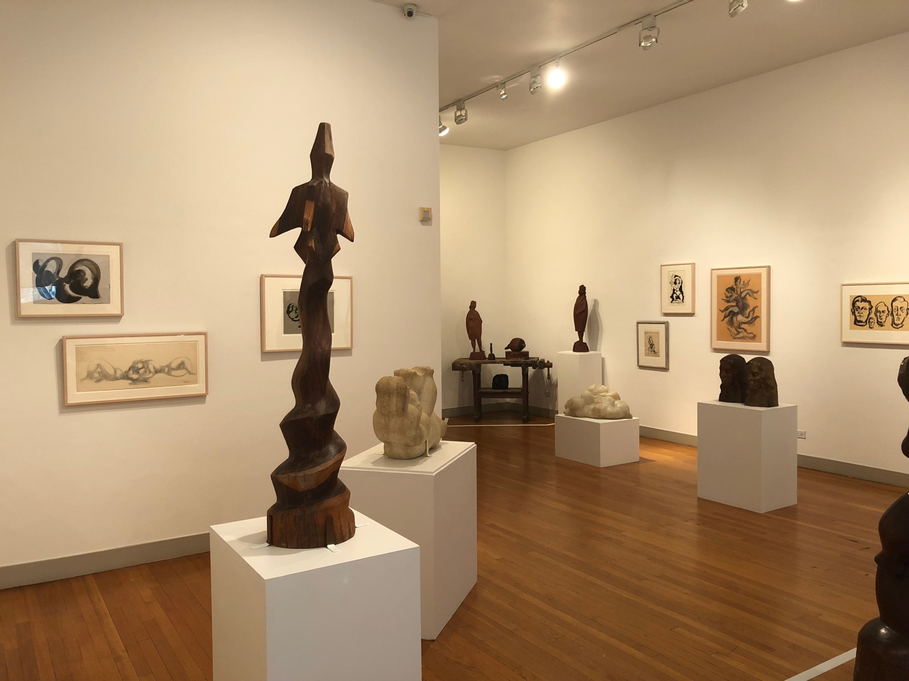Partial view of a gallery room and a hallway with white walls and medium brown flooring. There are many large sculpture on white rectangular mounts in organic shapes, in brown, white, and black. On the walls there are scatterings of framed sketches of the sculptures.