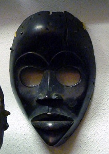 Mask made of wood and metal studs, replicating a human profile. The facial features include round cut-out eye cavities, enlarged lips, and a pointed chin.