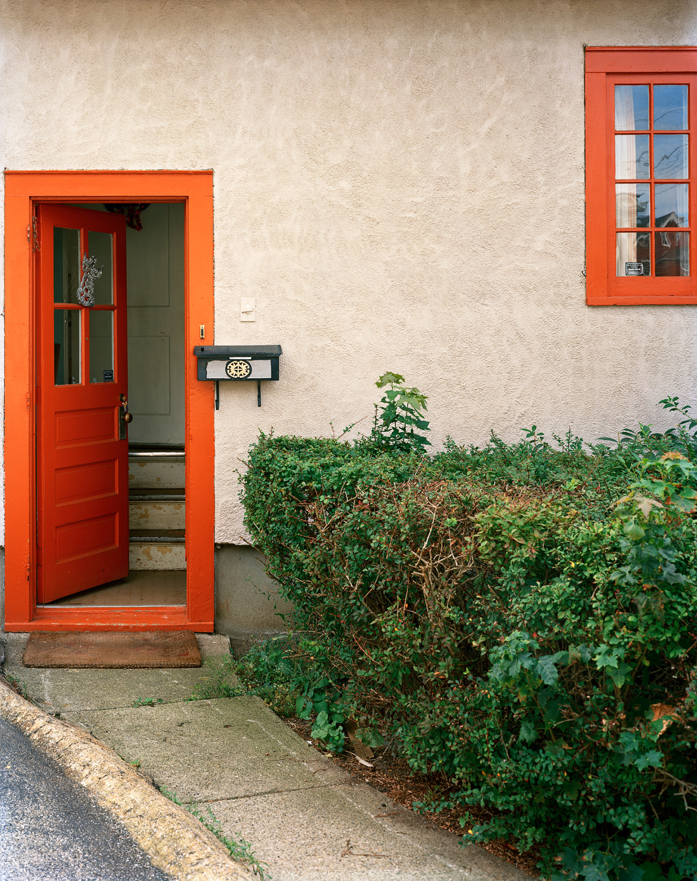Photograph of exterior of building with orange door and window and shrub, by Jade Doskow.
