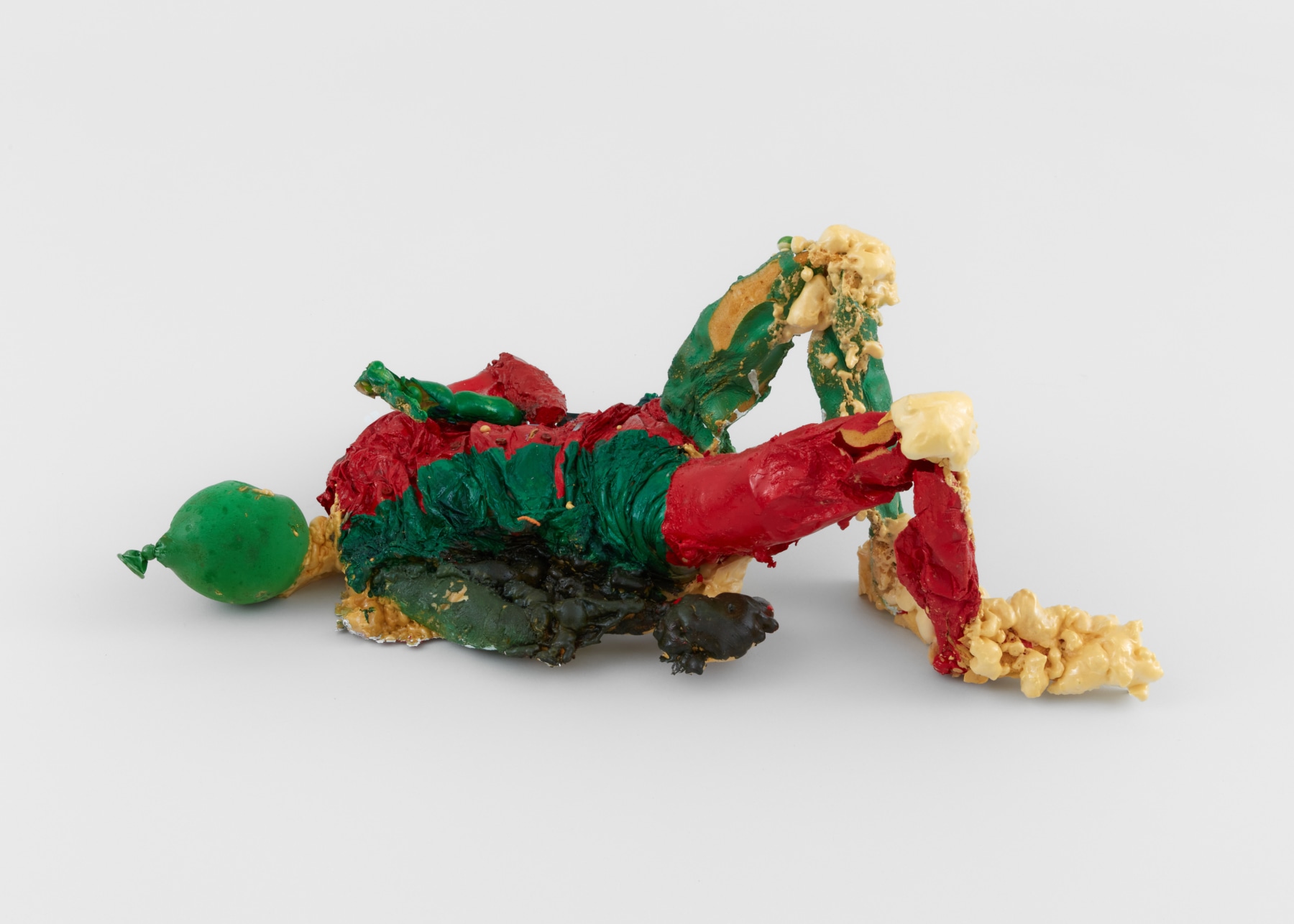 A free standing sculpture of a reclining figure painted red and green with a balloon for a head holds a pod of peas with spray foam bulging out from the knees and feet.