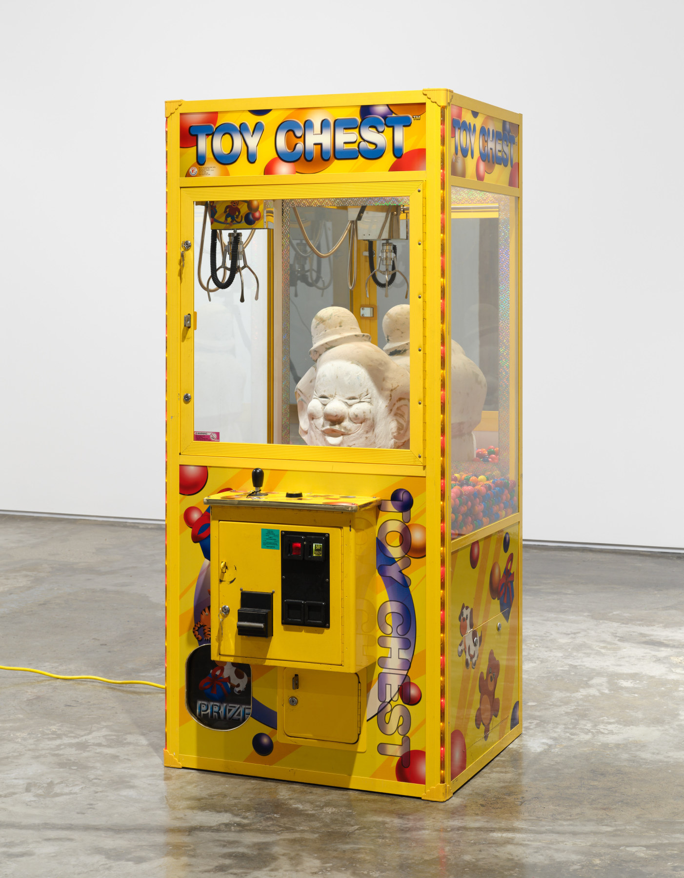 Anthony Olubunmi Akinbola's artowrk "Toy Chest". Yellow carnival game with large chalkware clown bust inside surrounded by gum balls. 70 x 31 1/2 x 33 1/4 in (177.8 x 80 x 84.4 cm), claw machine, candy, and chalkware, 2023.