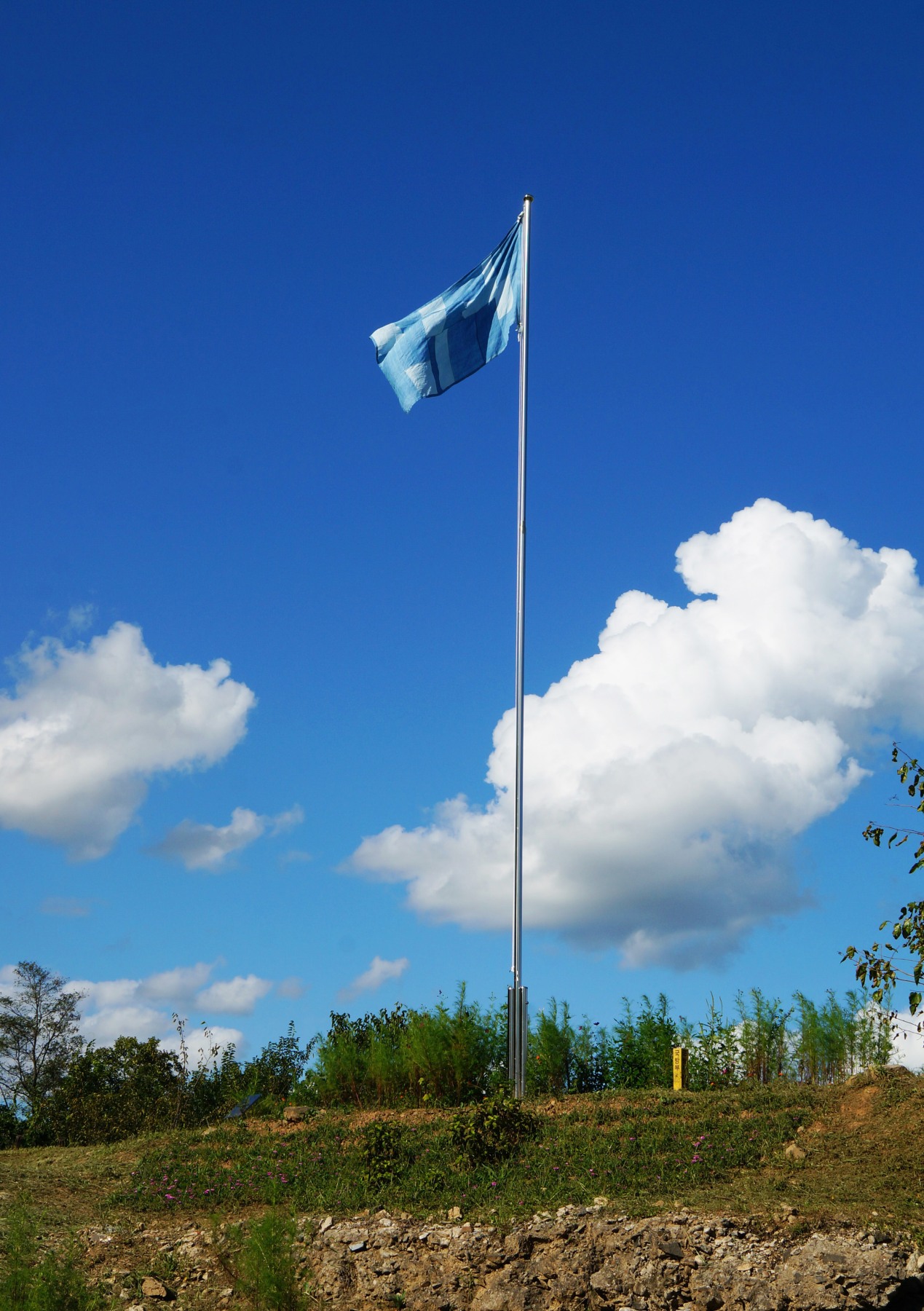 Sky blue flag on a flag pole stands under a clear sky and on soil with vegetation