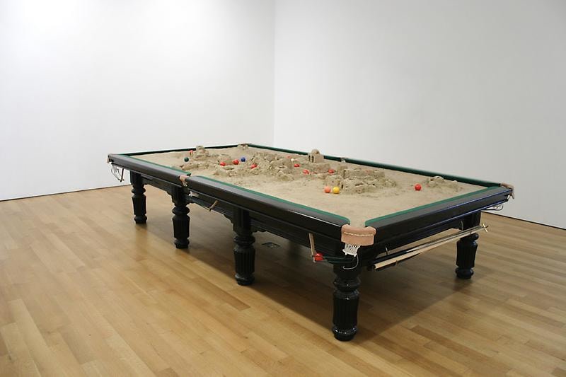 A pool table filled with sand with billiard balls scattered across it
