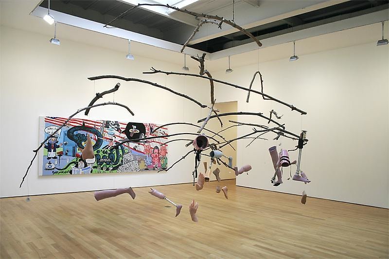 Installation view with a mobile of dangling prosthetic limbs hanging from branches