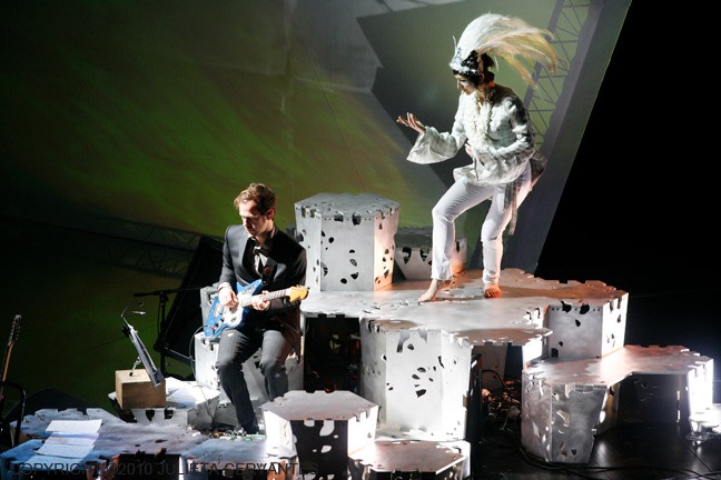 a woman dressed in white performing on top of a specially designed stage, looking down at a guitar player who is playing at a lowered portion of that stage