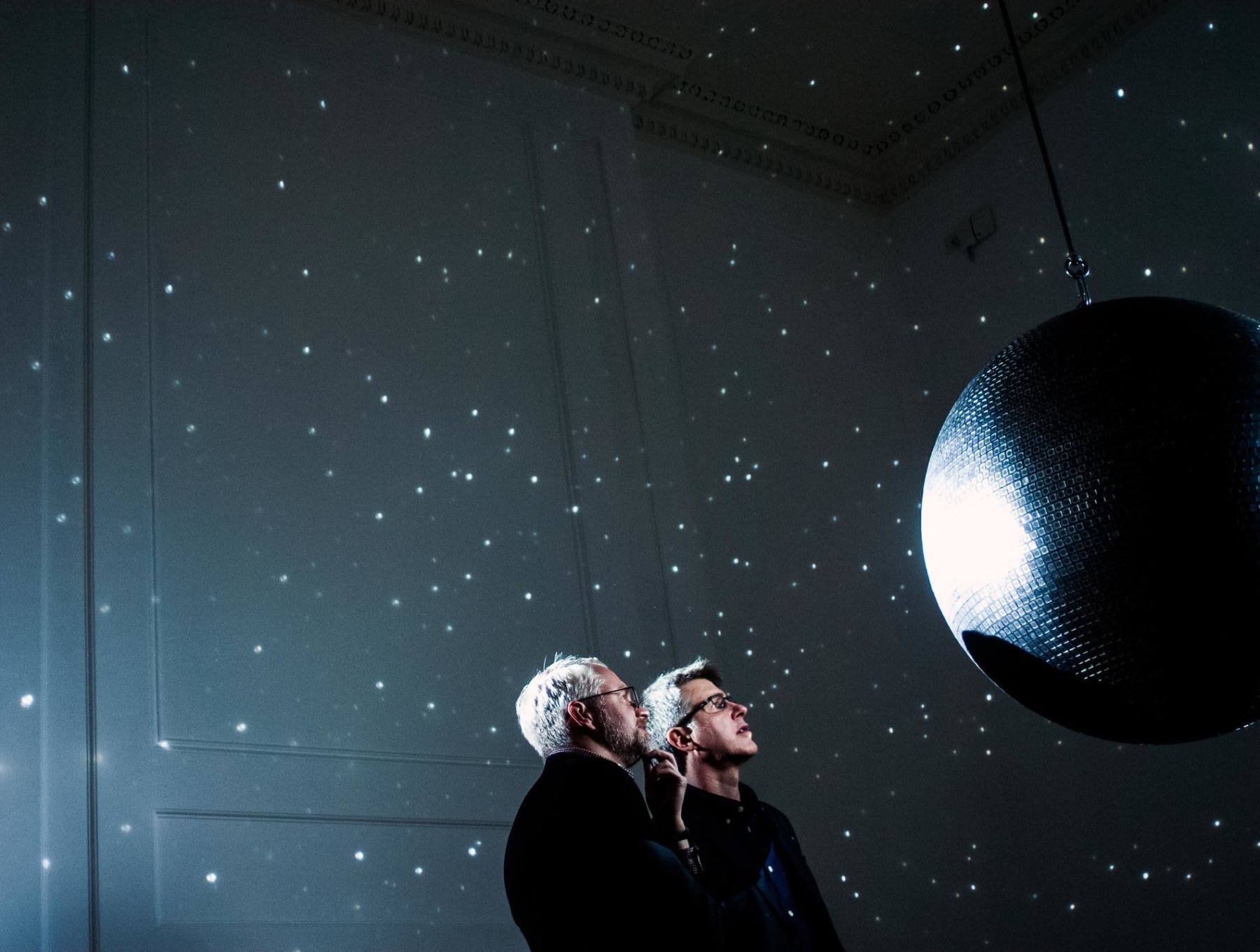 two men staring at a disco ball which reflect specks of light on a dark room