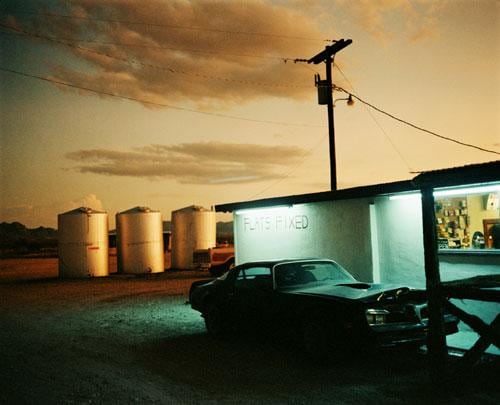 an abandoned car next to a white building with three storage tanks behind it