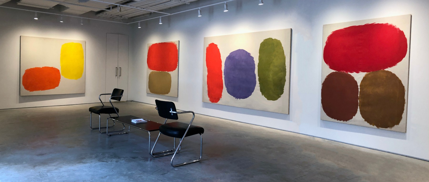 Four paintings by Ray Parker with colors of yellow, orange, umber, yellow ochre, violet, and red installed on a white wall.  Black table and two chairs are positioned in the middle of the room.