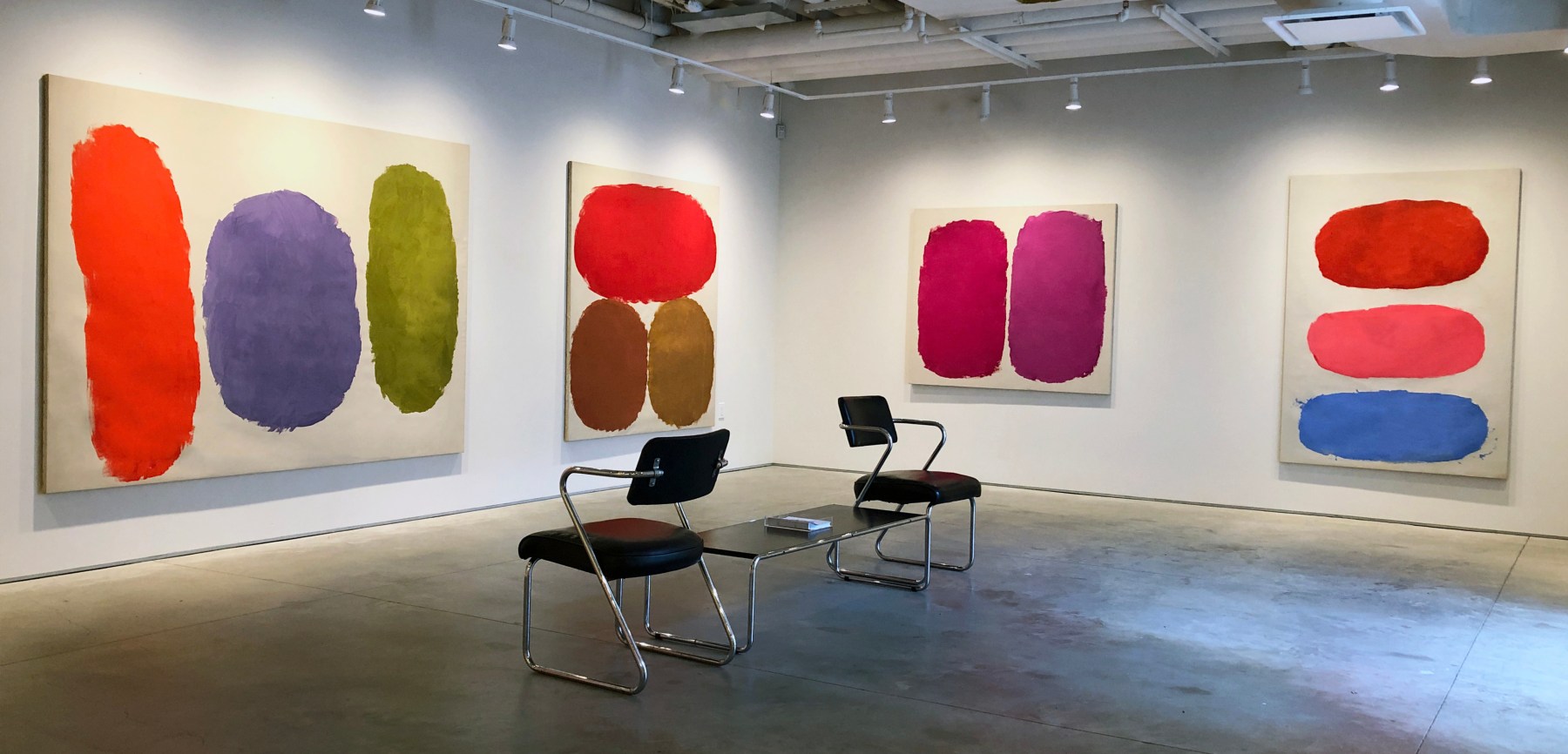 Four paintings by Ray Parker with colors of yellow, orange, umber, yellow ochre, violet, pink, blue, and red installed on a white wall.  Black table and two chairs are positioned in the middle of the room.