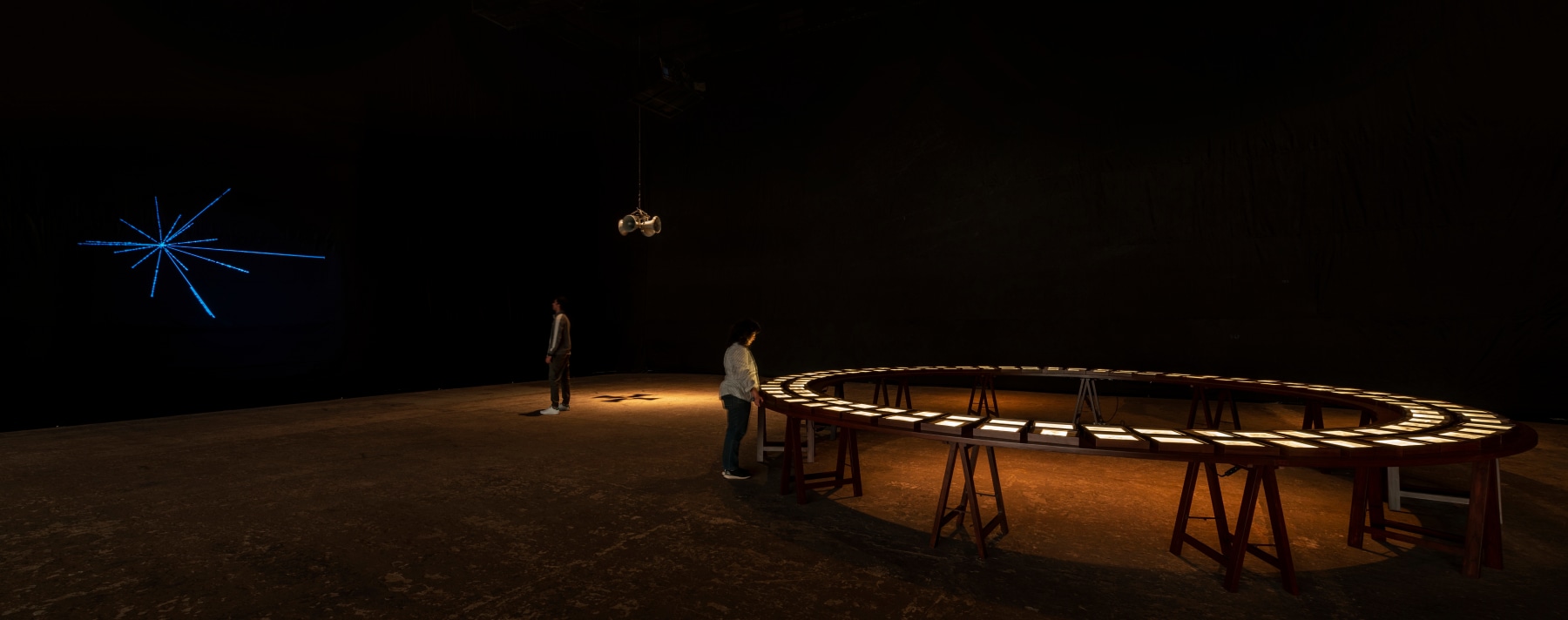 Installation view of Jitish Kallat's "Covering Letter (terranum nuncius)". On left is a blue light sculpture, a speaker hangs in the middle, and there is a large round table on the right.