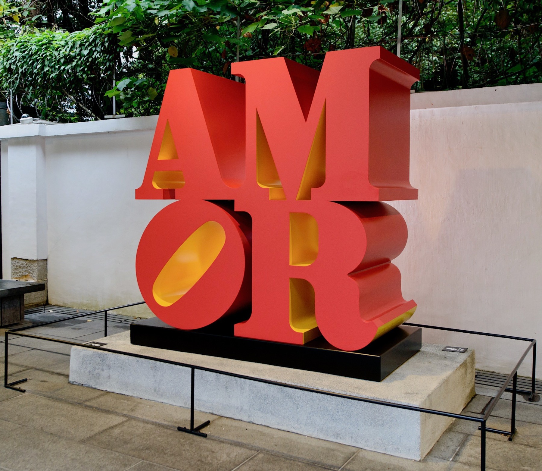 Installation view of the red and yellow polychrome aluminum sculpture AMOR, with the letters A and M stacked on top of a titled letter O and the letter R.