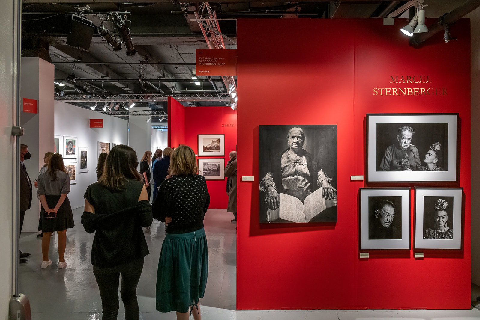 People walk through the aisles of The Photography Show near a bright red wall with black and white photos.