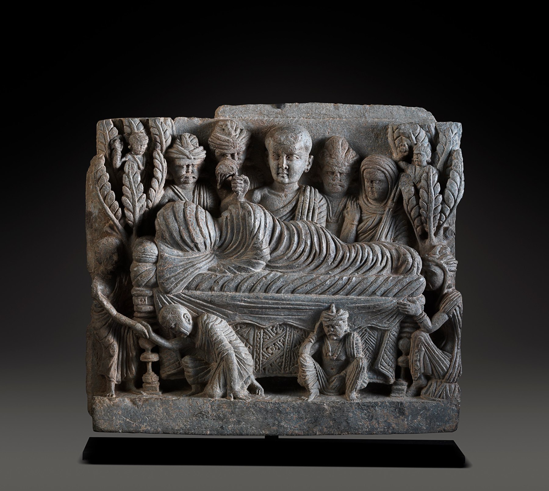 This relief shows the parinirvana of the Buddha in a rather unusual manner.  The teacher, on his death bed, is depicted at the center of the relief.  His head and body are completely swathed in cloth, and his face is not visible.