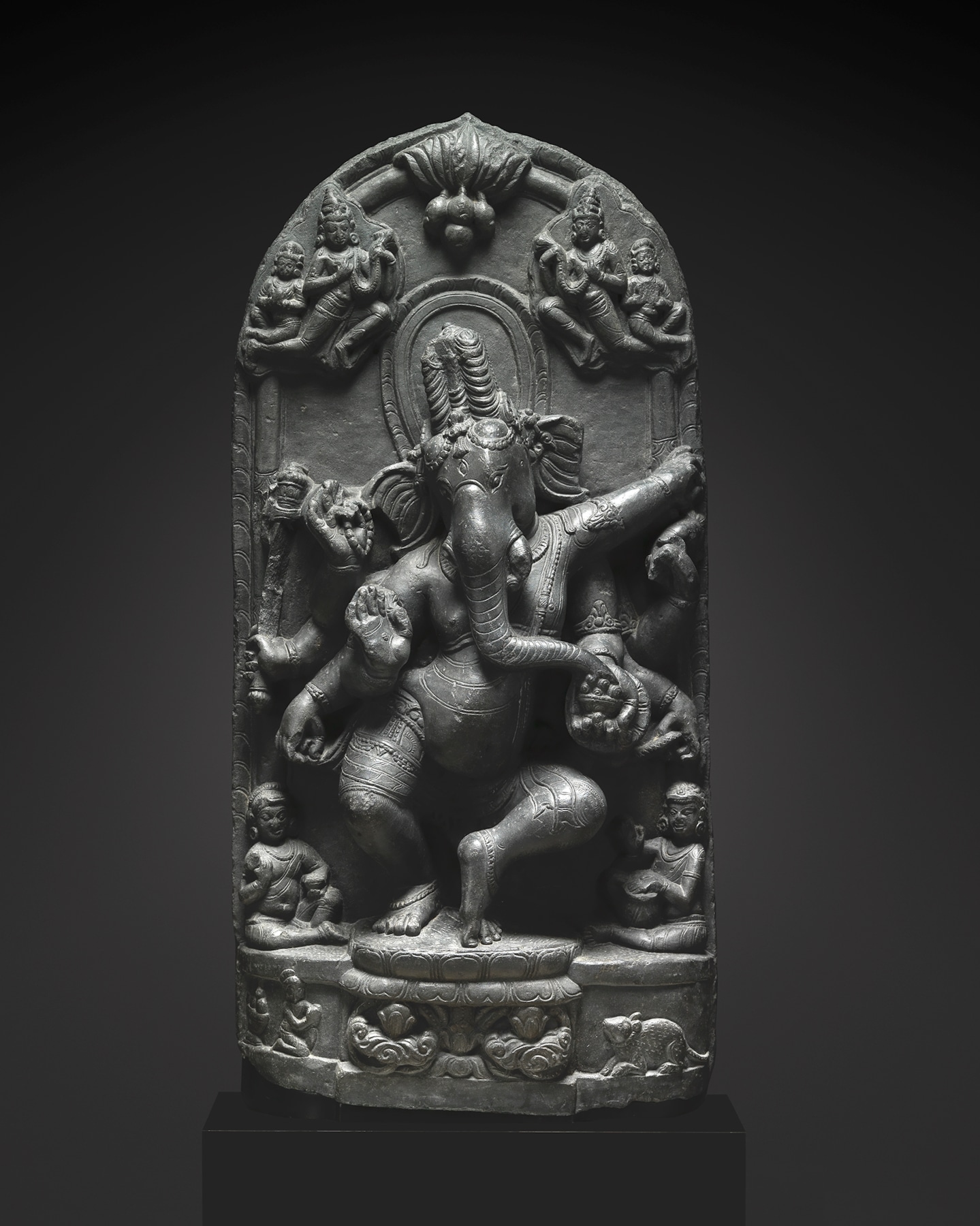 Watched by two cloud-borne celestial garland bearers, eight-armed elephant-headed Ganesha dances in a lively attitude on a lotus flanked by his recumbent rat vehicle and a kneeling donor figure.