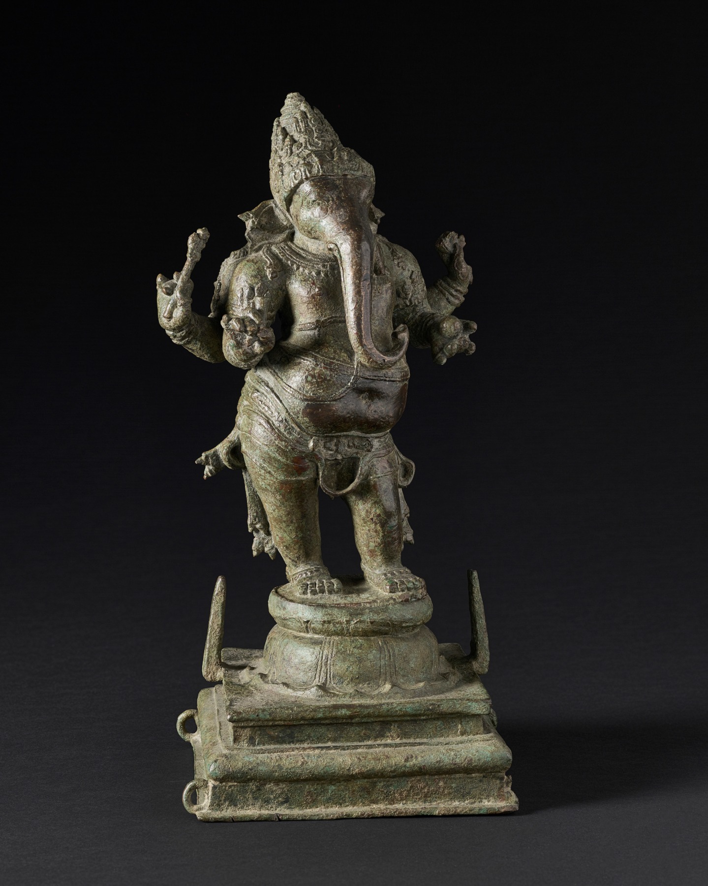 In this charming sculpture, Ganesha stands in bulging contrapposto atop a circular lotus supported by a molded square base with lugs on the sides and vertical pegs. 