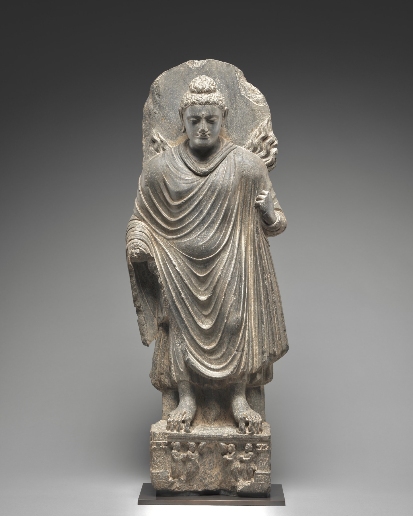 The Buddha is standing in a subtle tribhanga pose atop a rectangular pedestal with an inset panel depicting devotees.
