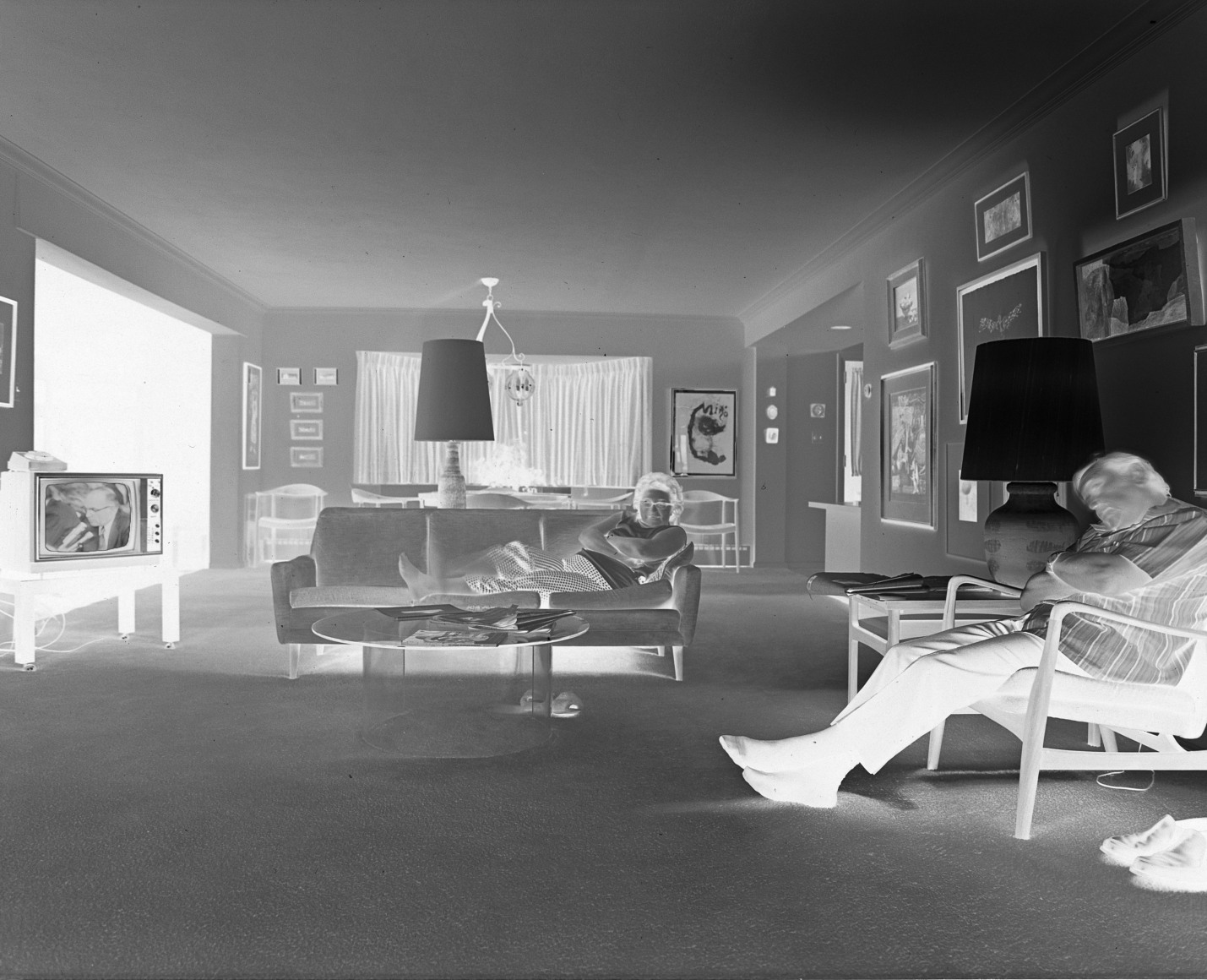 Negative printed photo of a couple sitting in a midcentury living room with the TV on. The man sleeps in a char while the woman looks at the camera.