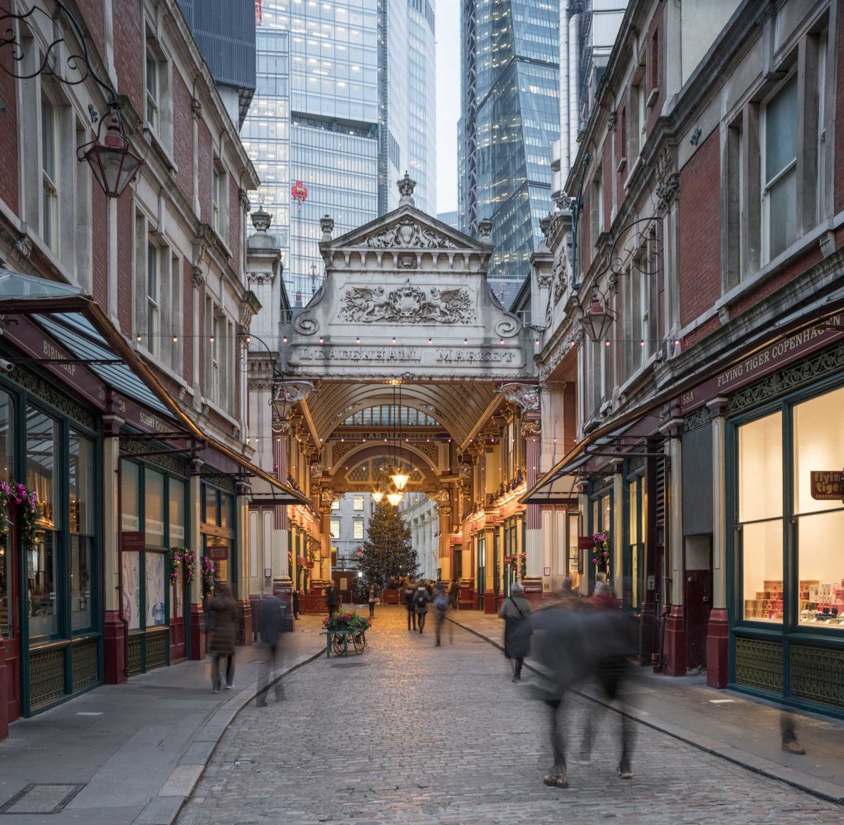 Color long exposure photo of holiday shoppers at an historic outdoor market in London, a Christmas tree can be seen under a Victorian arch in the center background.