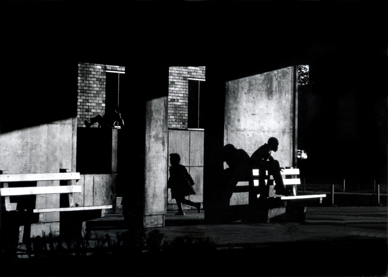 Shadowy black and white image showing young black children playing in an urban area, one sits on a bench, another in a window, one runs through a door.