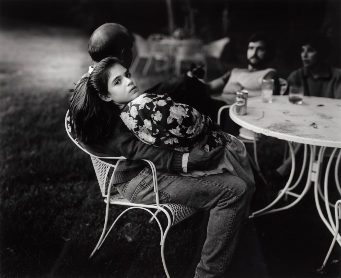 Black and white photo of an adolescent girl sitting on an adults lap at an outdoor table, looking back at the camera.