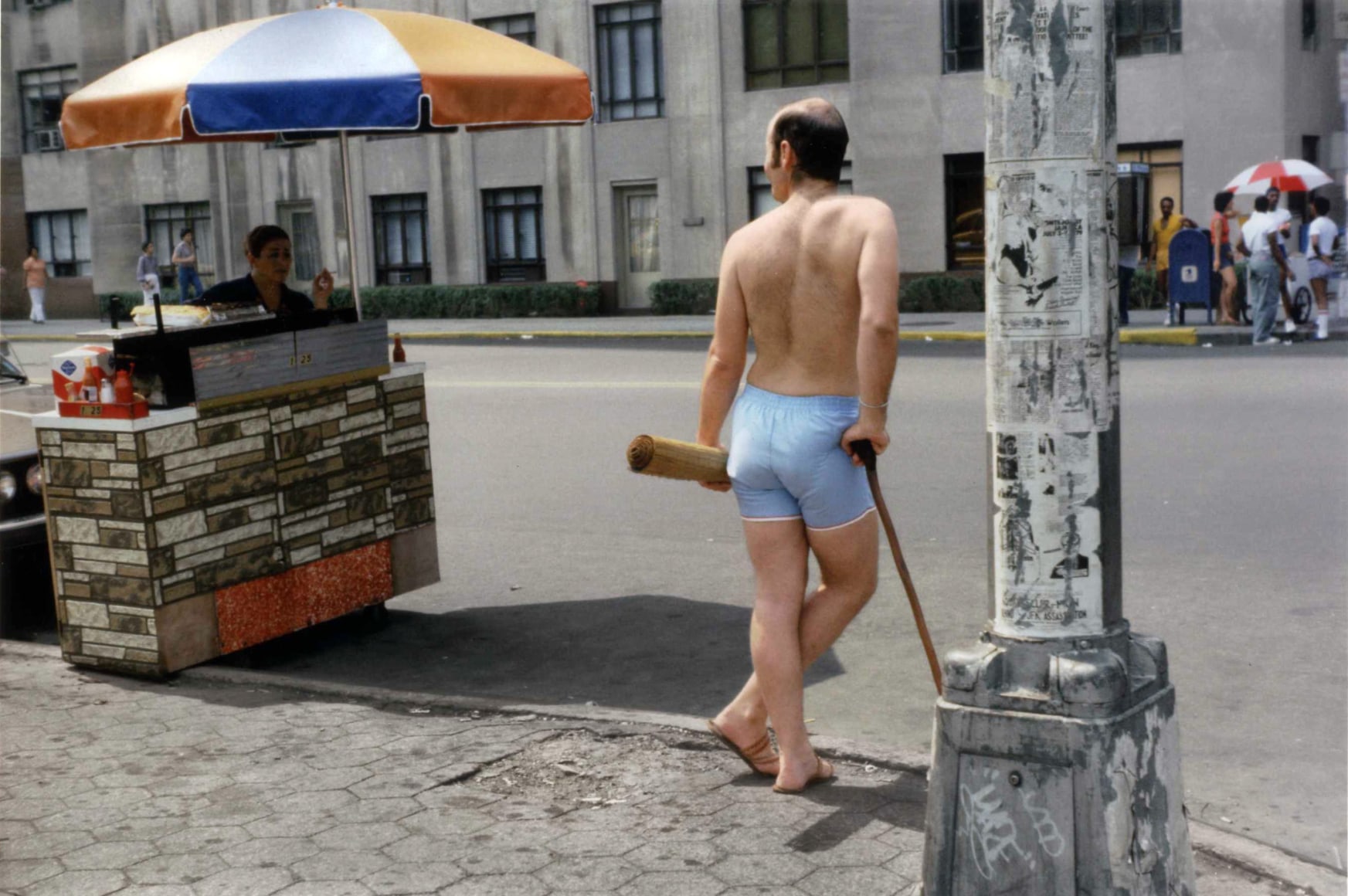 Color photograph of a man in athletic shorts and no shirt leaning on a cane with a rolled mat under his arm at a CYC intersection.