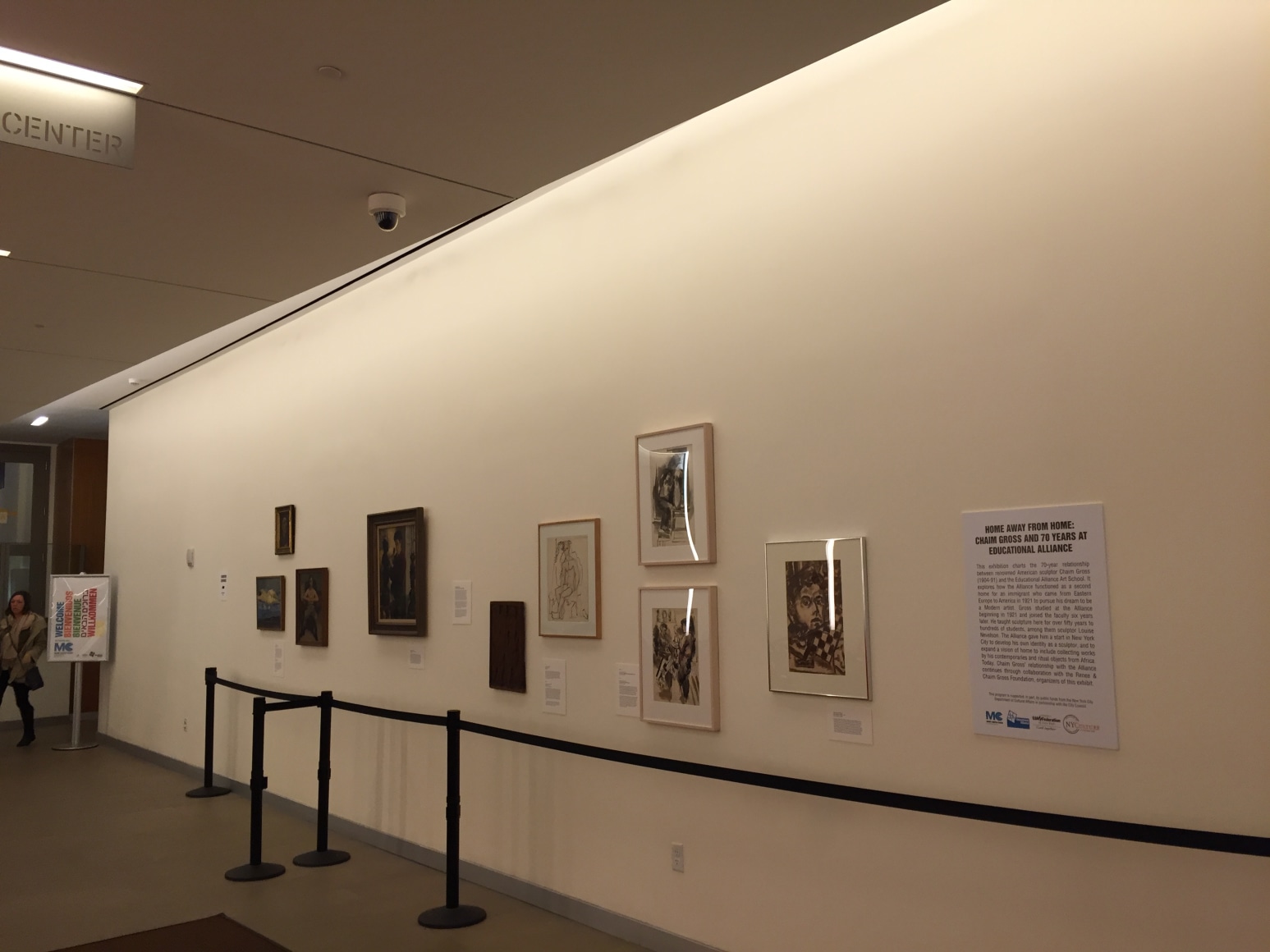 Long white gallery wall going to the left with grey flooring beneath it. At the far left of us an adult wearing a winter coat and black rain boots walks to to the left. On the gallery wall there are two distinct clusters of framed art works, the cluster furthest away from us has four dark colored pieces, one large and the other three small, all in dark brown frames. The cluster closest to us has four similar sized medium pieces on white paper with cream frames. Between both of the clusters there is a black painted, medium sized piece of canvas or wood.