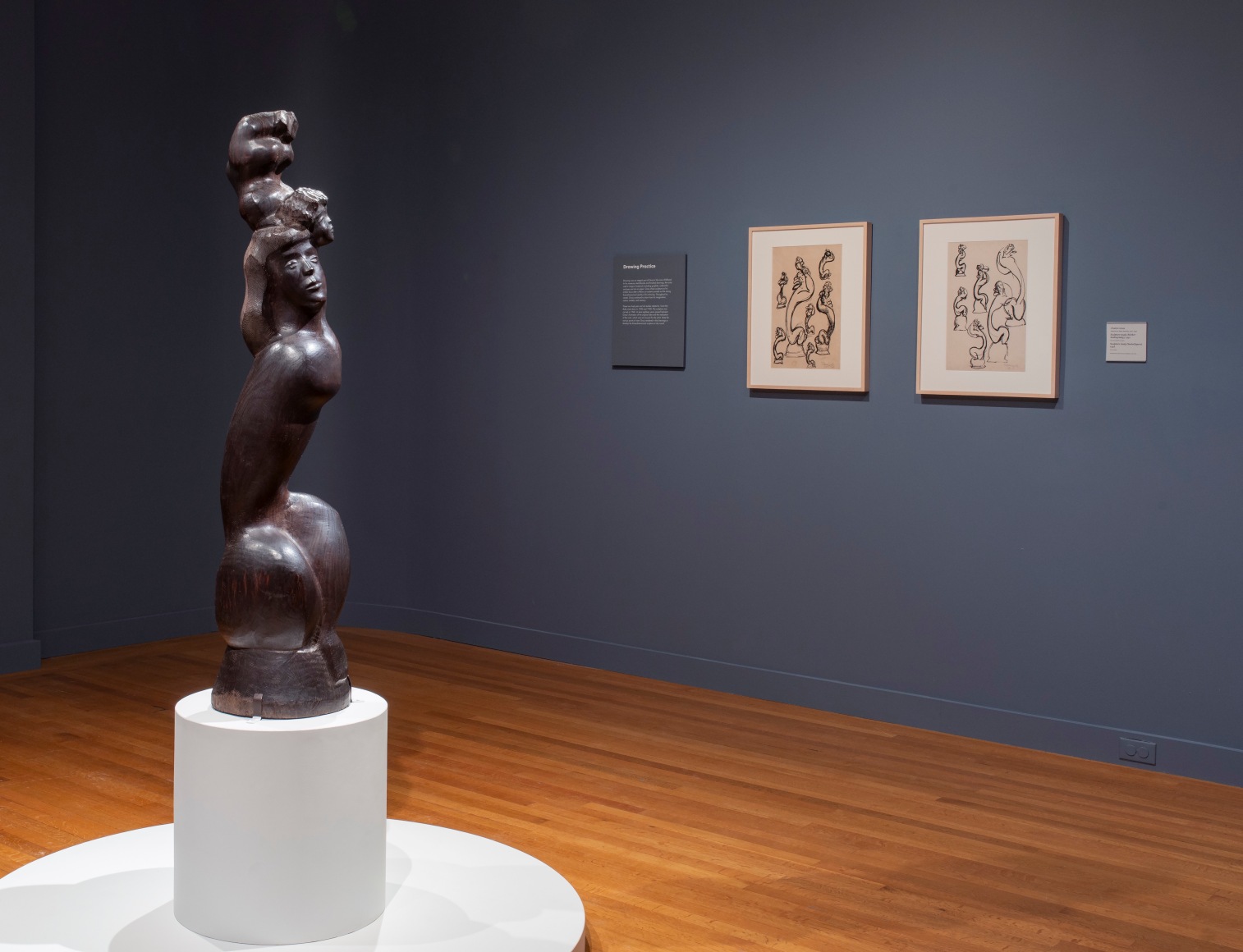 Photo of a wooden sculpture, I LOVE MY BABY, 1948, depicting a woman with long hair, crouched so that her legs are drawn close to her tall torso. She is supporting the figure of a baby, whose head touches hers but whose body is raised above its head. To the right of the sculpture are two framed ink sculpture studies for the same sculpture. The walls are faded navy.
