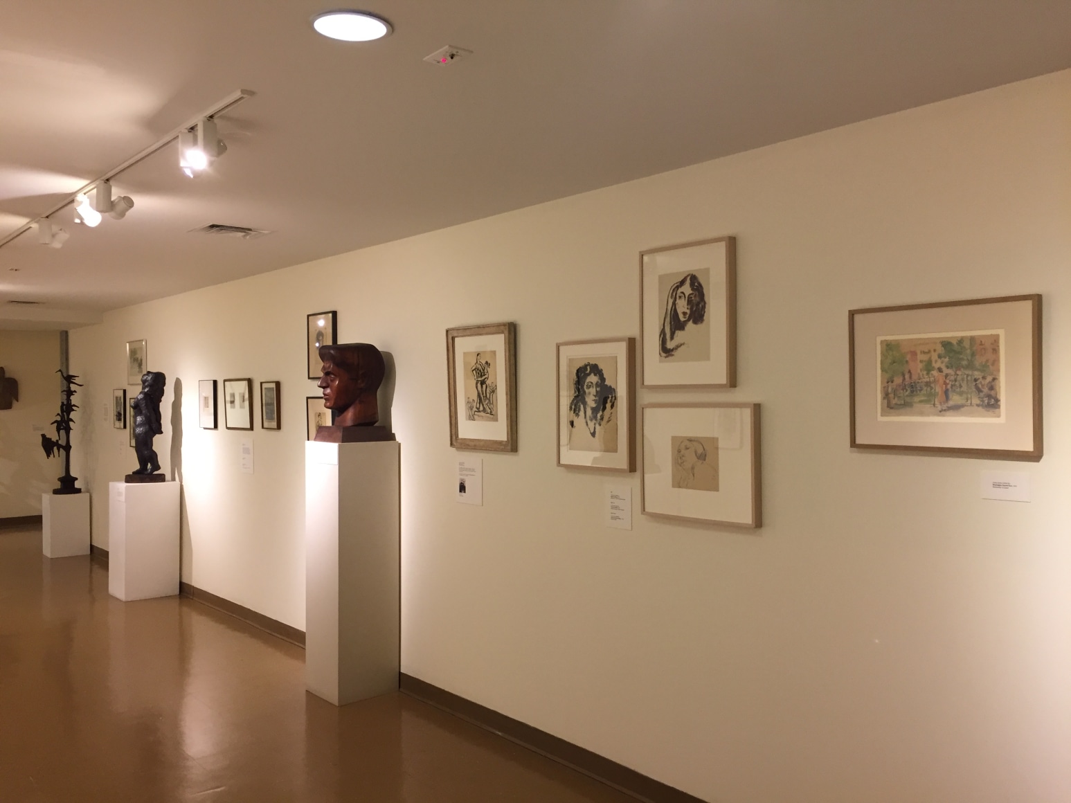 White gallery wall going diagonally towards the left with medium brown flooring beneath it. On the right closest to us there is five medium sized pieces of art work in beige frames. They all are depictions of people in black and white except the one closest to us, which illustrates a colorful scene of a park. Next to the art works to the left is a mid size, medium brown bust of a man, to the left of that sculpture are more pieces of artwork hung up and two medium sized black sculptures of a human figure and a tree with birds on it, also with art hung between them.