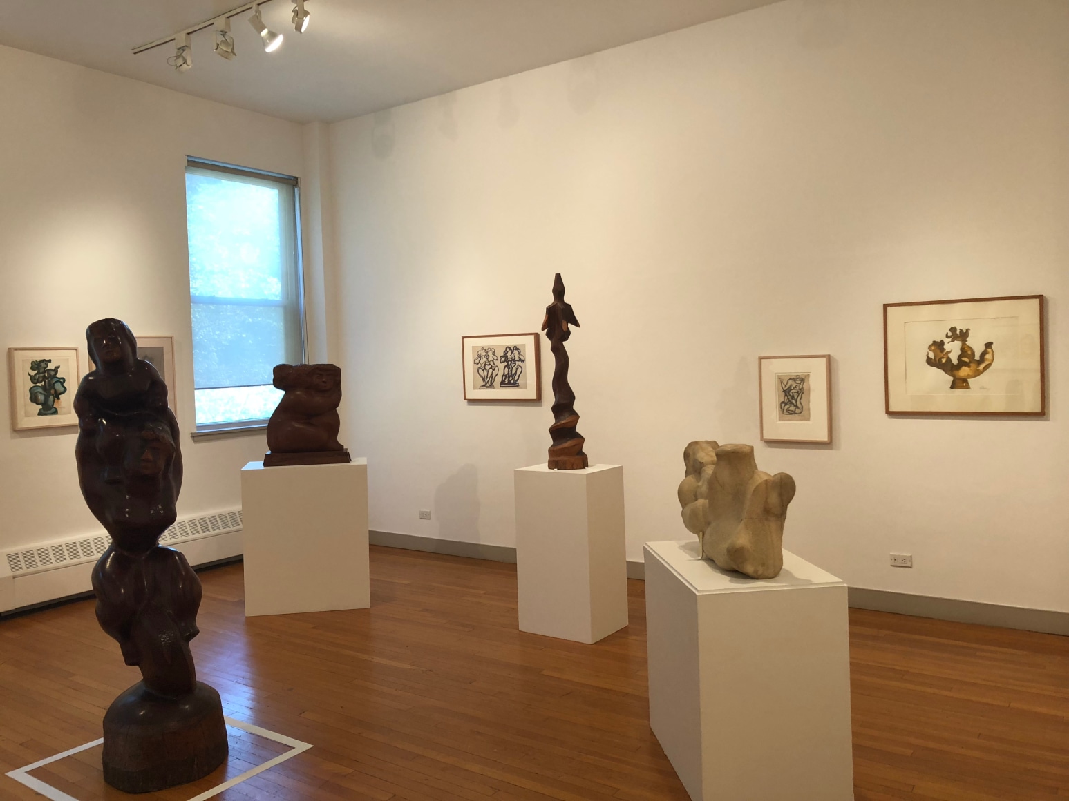 Photo of a gallery with two white walls intersecting at the left and medium brown, wood flooring. There are sculptures from left to right of: a tall, thin, black composition of bulbous forms, a large rectangular woman, a thin, medium brown bird like shape, and an off white, short sculpture. On the walls there are framed artworks depicting sketches of the sculptures.
