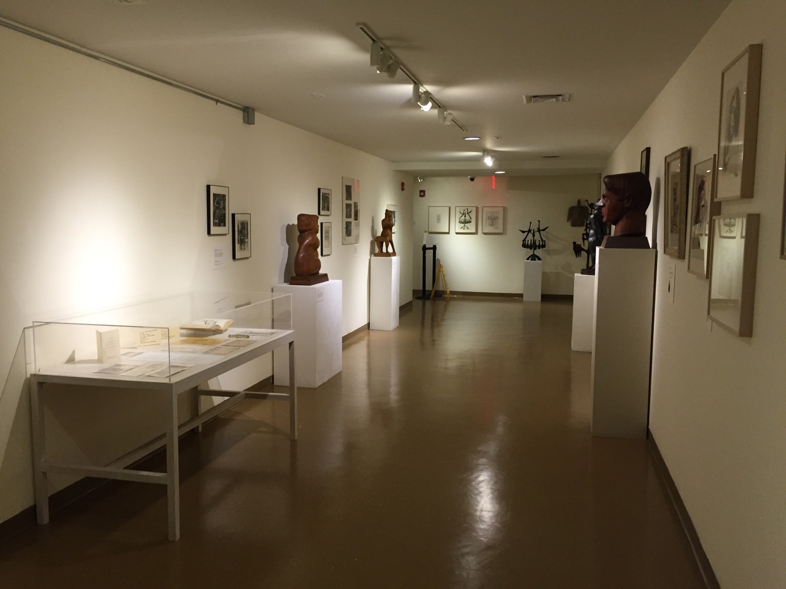 Hallway of a gallery with white walls, medium brown floors, and a low white ceiling. On the left wall closest to us is a glass box mounted on a white table with papers and a book inside. Next to it on the left wall towards the right is a medium brown, mid sized sculpture of a vaguely humanoid, organic shape, and to the right of that piece is a unidentifiable animal like, organic form. There are four medium sized framed artworks and one large piece between and around these sculptures. On the right wall there is a large, medium brown bust of a man and a piece of a black unidentifiable sculpture is visible. Closest to us on the right are the sides of four artworks in beige frames. On the back wall there is a black sculpture of a tree with birds on it and three mid sized artworks hung up to the left of it.