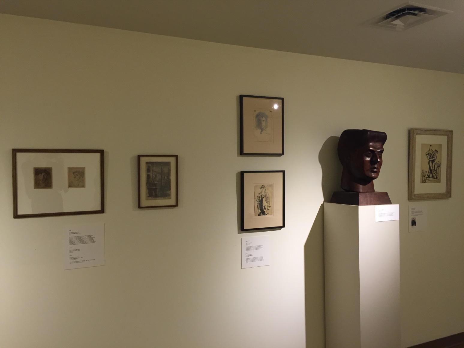 White gallery wall with a low ceiling with five pieces of framed artwork on it and a medium brown, mid sized bust of a man's head on the right. The four pieces of artwork to the left of the sculpture are in dark brown frames and depict sketches of people and the illustration in the center appears to be of buildings. The art work on the right side of the sculpture is in a beige frame and shows two people intertwined.