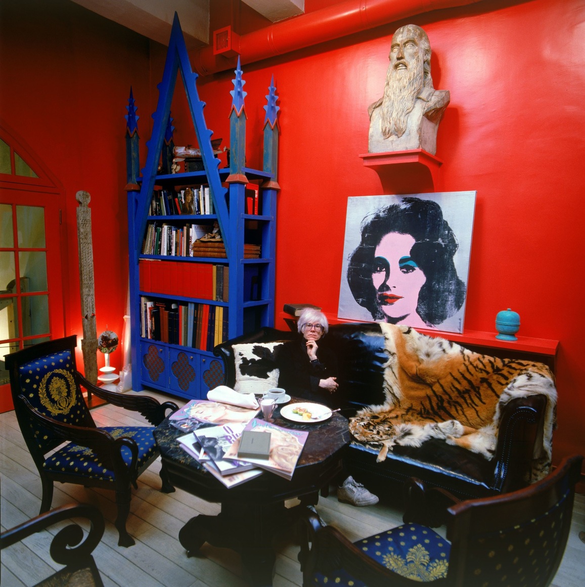 Tseng Kwong Chi, Andy Warhol, New York, with Liz Taylor&rsquo;s portrait in Fred Hughes&rsquo; red studio, 1986. Chromogenic print, image: 19 1/2 x 19 1/2 inches, frame: 26 1/2 x 26 1/2 inches.