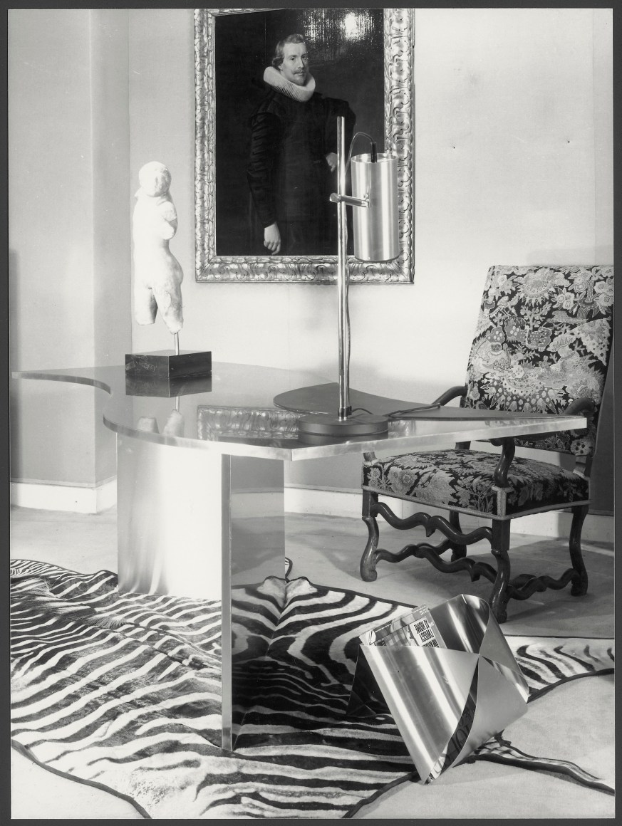 Black and white photo of a metal wave shaped desk, metal envelope magazine holder, and traditional chair atop a zebra rug