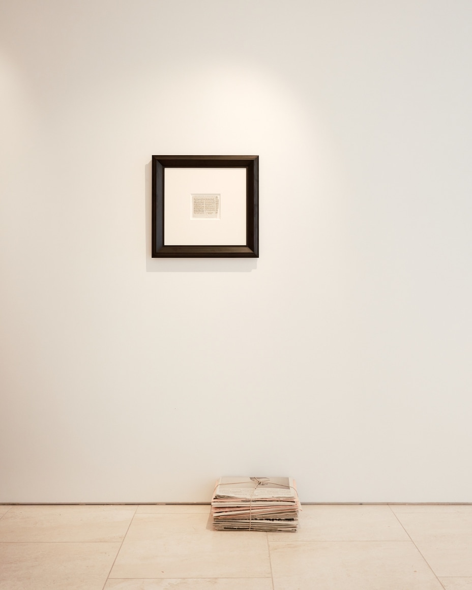 a newspaper clipping with teared edges hangs in a frame above a stack of newspapers in a gallery