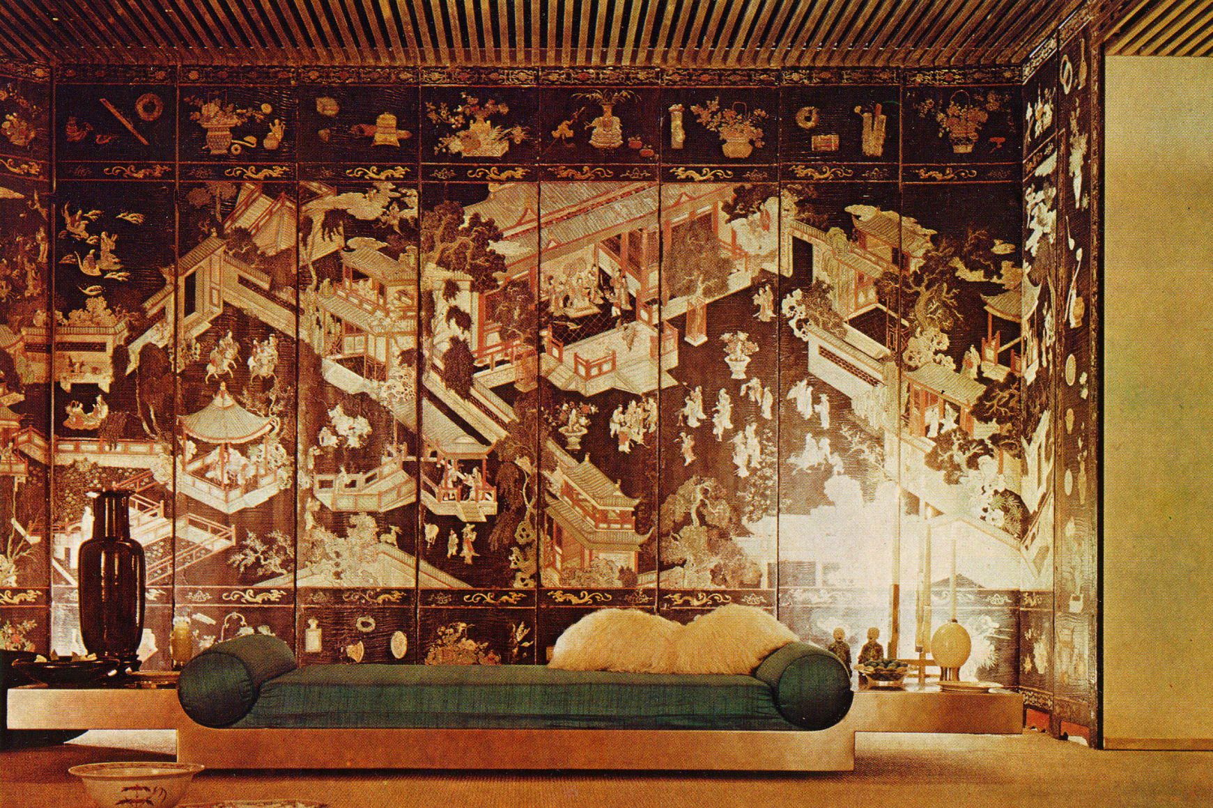 Archival photo of a metal daybed with extended table arms and upholstered cushion and round pillows in front of a large tapestry