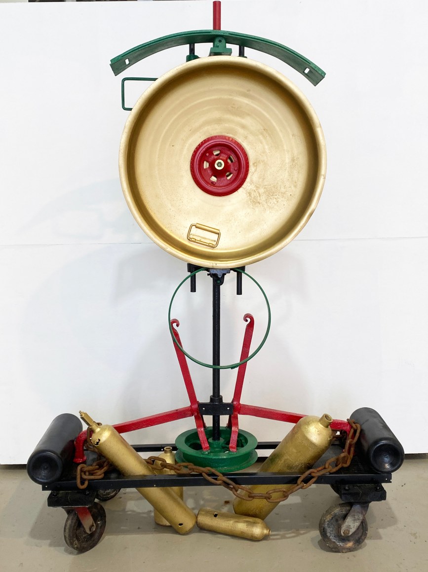 Assemblage sculpture made with found objects such as canisters, lids, wood, wheels. Colors are gold, red, green and black