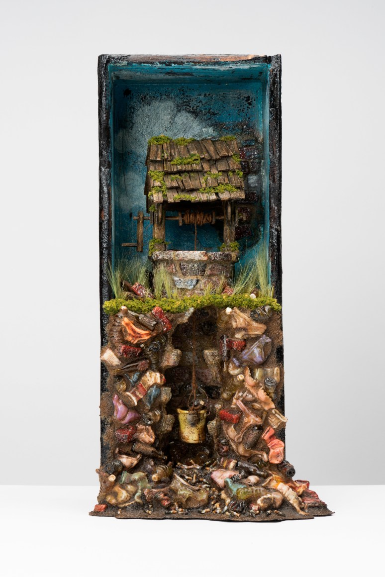 Box sculpture of a well on a grassy surface sitting a top a pile of used clothes and trash