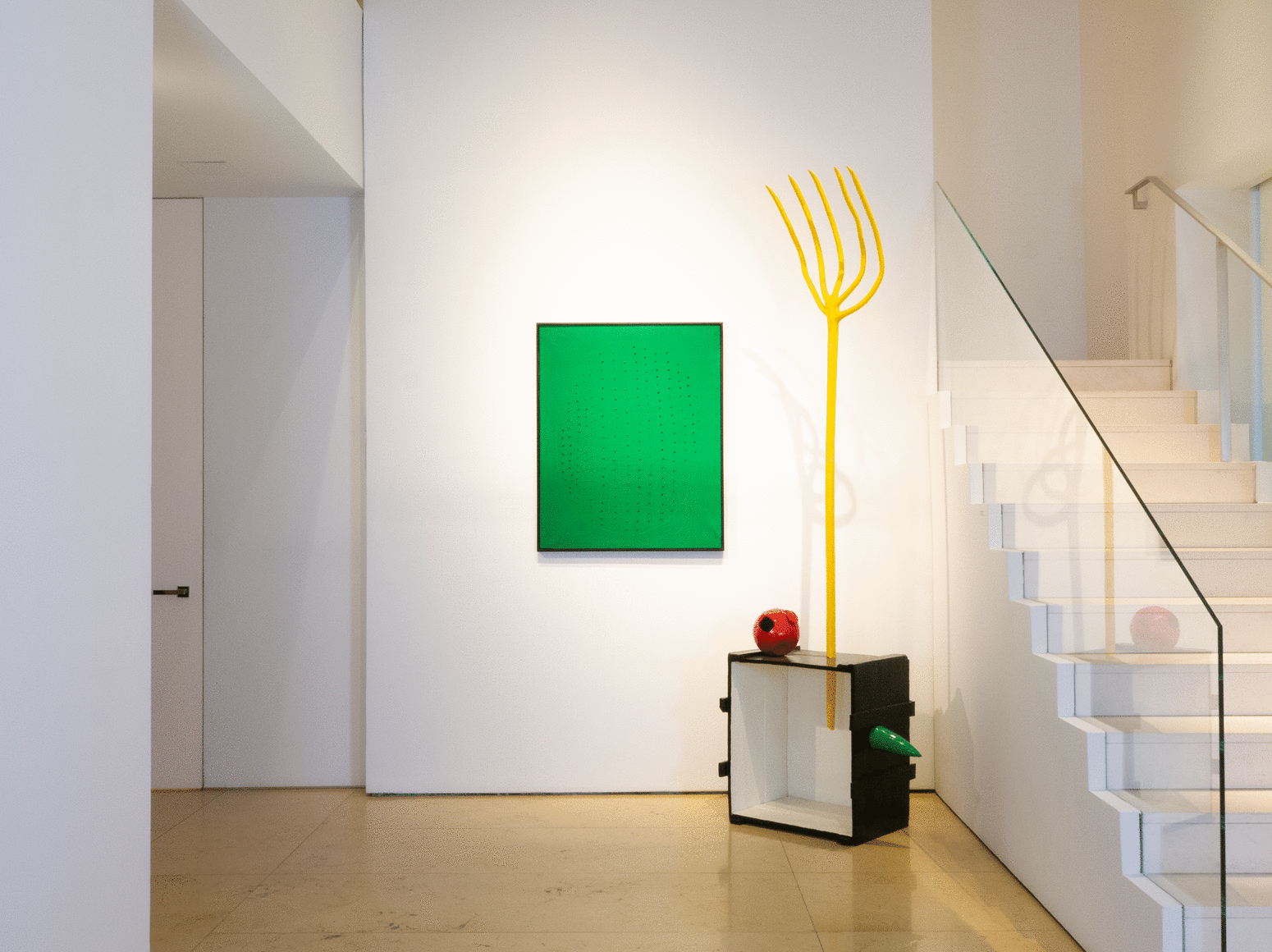 theInstallation view of Masterworks. This photo feature a green painting by Lucio Fontana pierced with holes contained in the shape of a circle. Next to the Fontana painting on the right is a large scale sculpture by Joan Miro, in an assemblage manner made of found objects later poured in bronze and painted over, we can see a box standing on one of its side, pierced on the top by a tall yellow fork. There is also a red ball seating on the box.