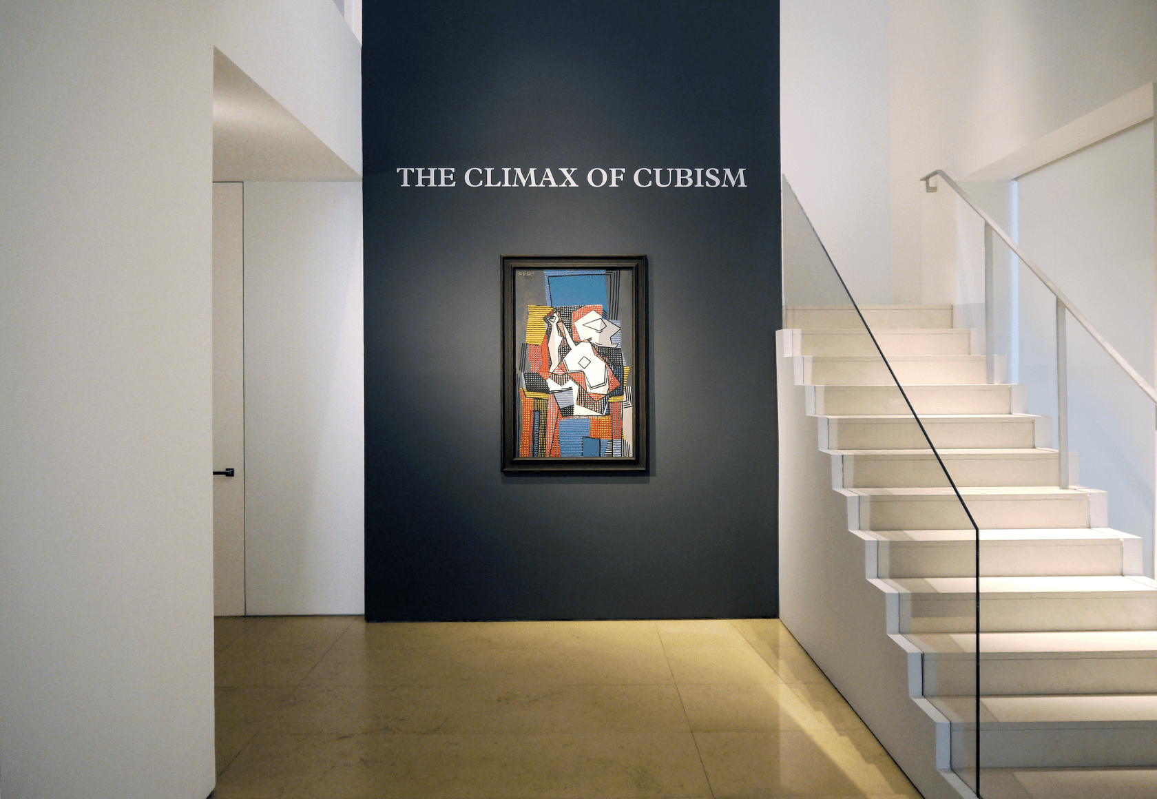 Installation view of the lobby entrance which features the title in white vinyl over a grey wall and a Picasso painting called Bouteille, Guitare Compotier, made in 1922. The painting represents a white guitare depicted in a cubic manner over many juxtaposed grids executed with various colors, red, yellow, blue, black. The overlapping grids create a vibrant composition.