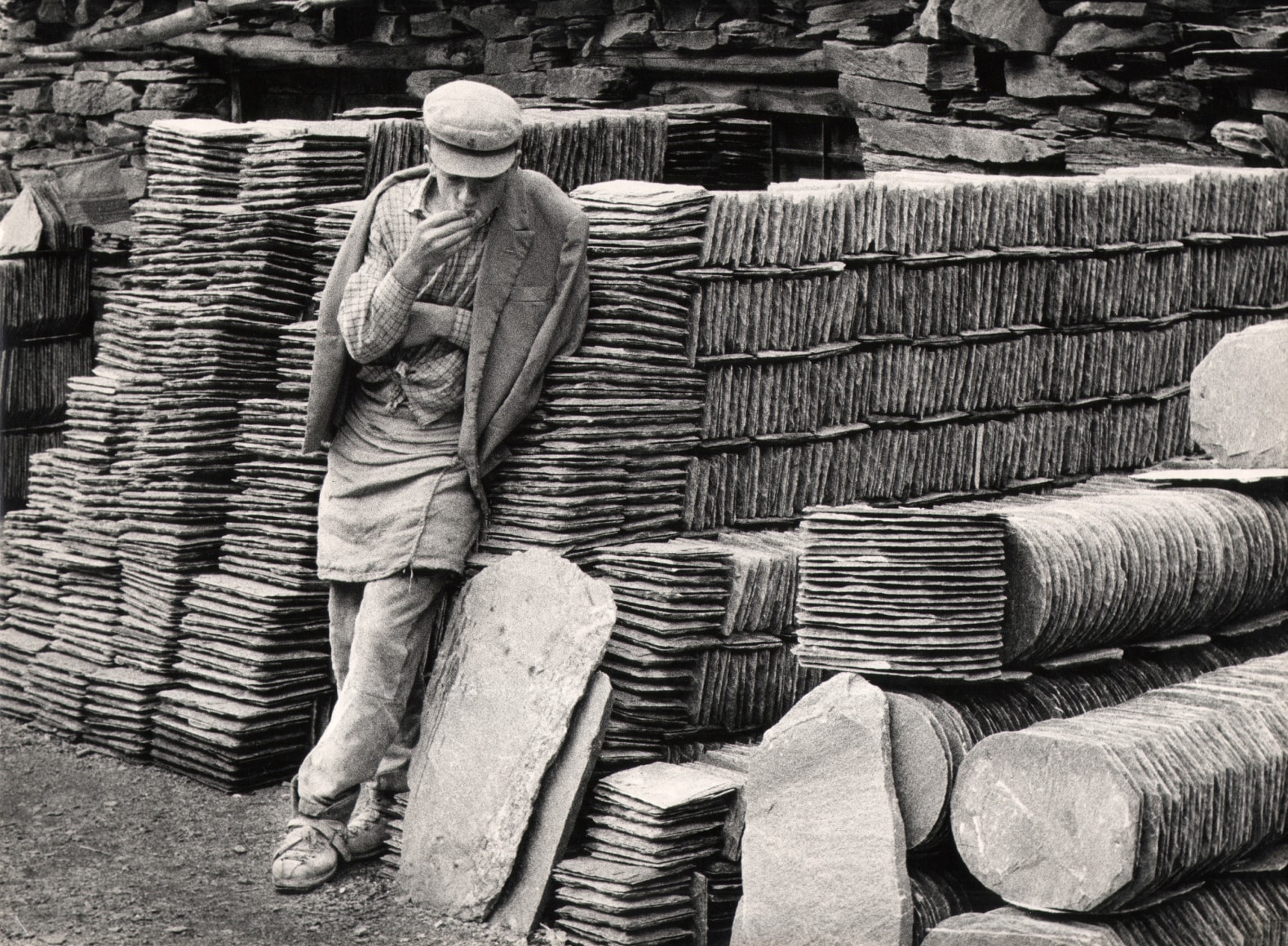 Mario Finocchiaro, Pietra Ardesia, Cava di Branzi, ​1950. A quarryman stands smoking a cigarette, leaning against stacks of slate.