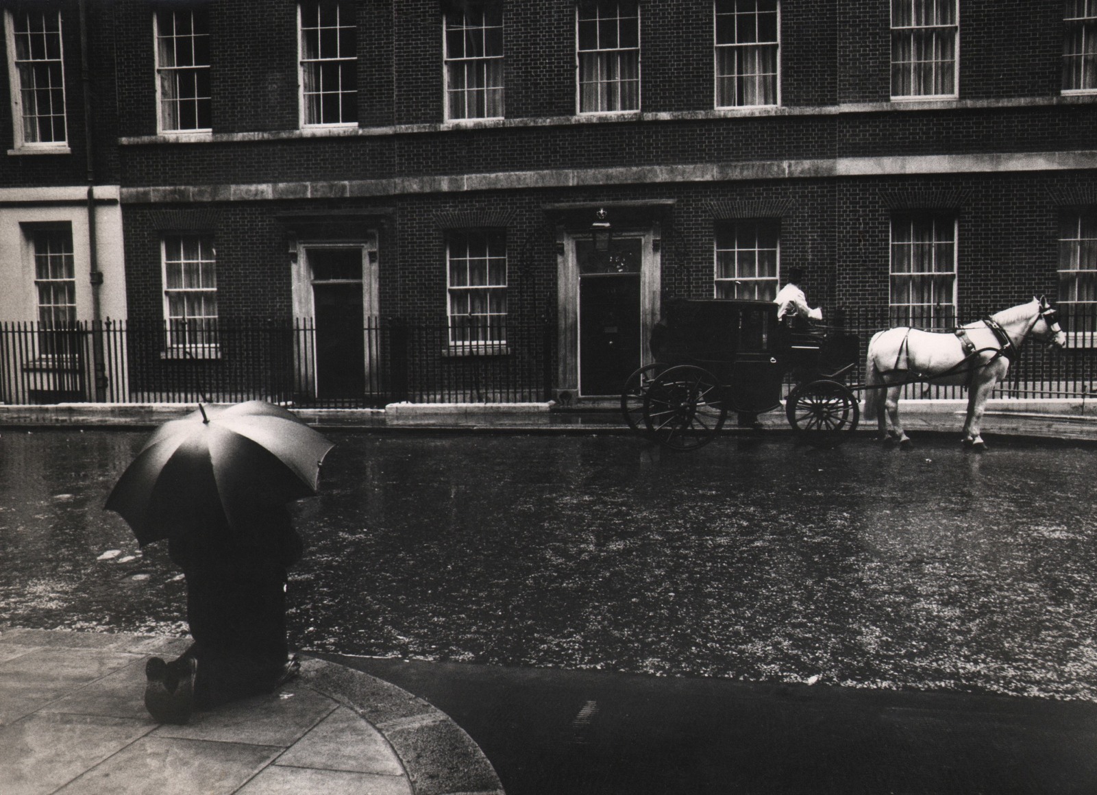 31. David Newell Smith, The man in Downing Street was praying for Mr. Wilson who had just become the Prime Minister, ​c. 1966. A figure with an umbrella kneels on the lower left of the frame across the street from a horse-drawn carriage.