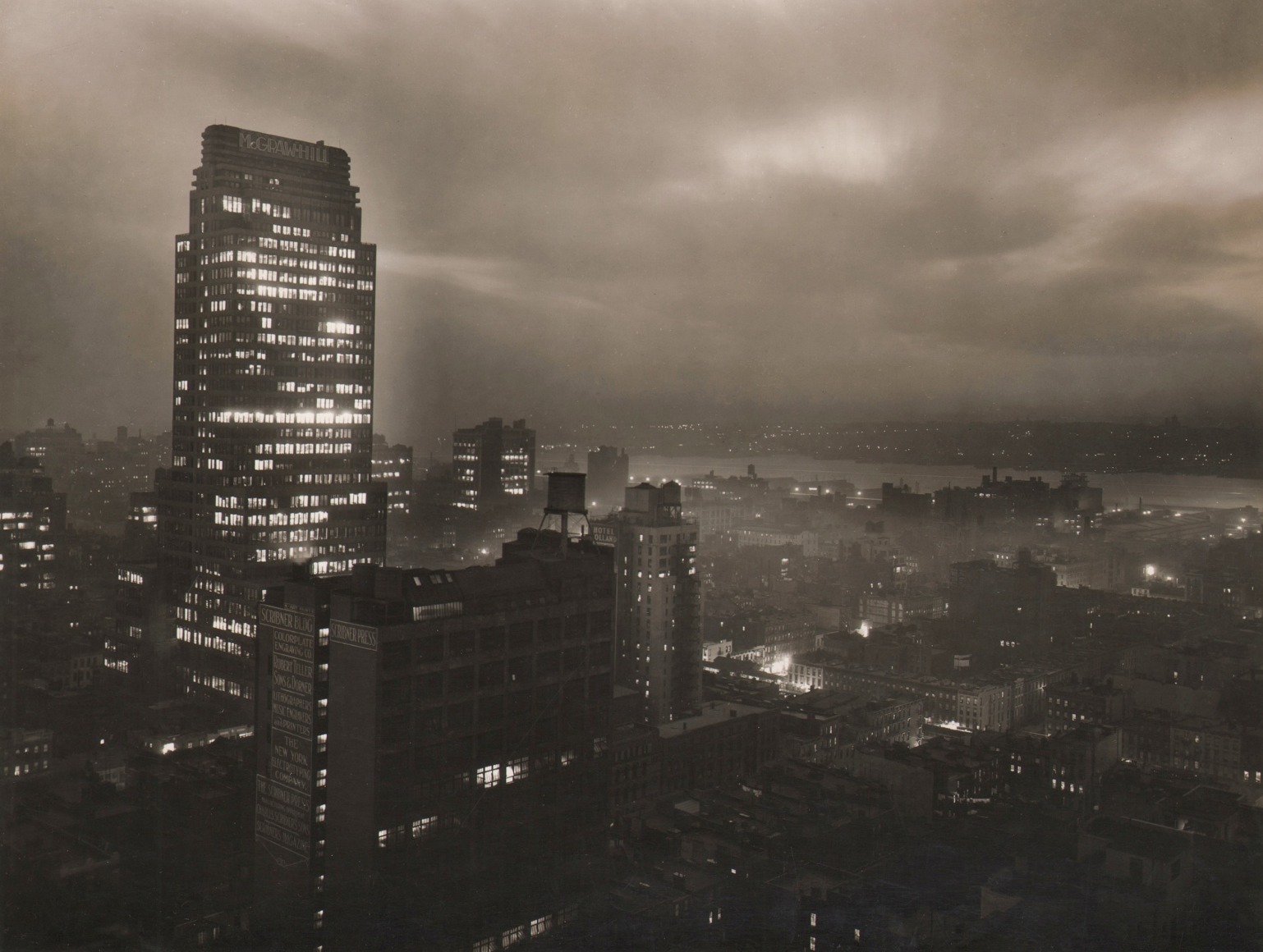 Paul J. Woolf, McGraw Hill Building &amp; Hudson River from Roof of the Hotel Lincoln, ​c. 1935. Night time cityscape with tell McGraw Hill Building in center left of the frame beneath overcast sky.