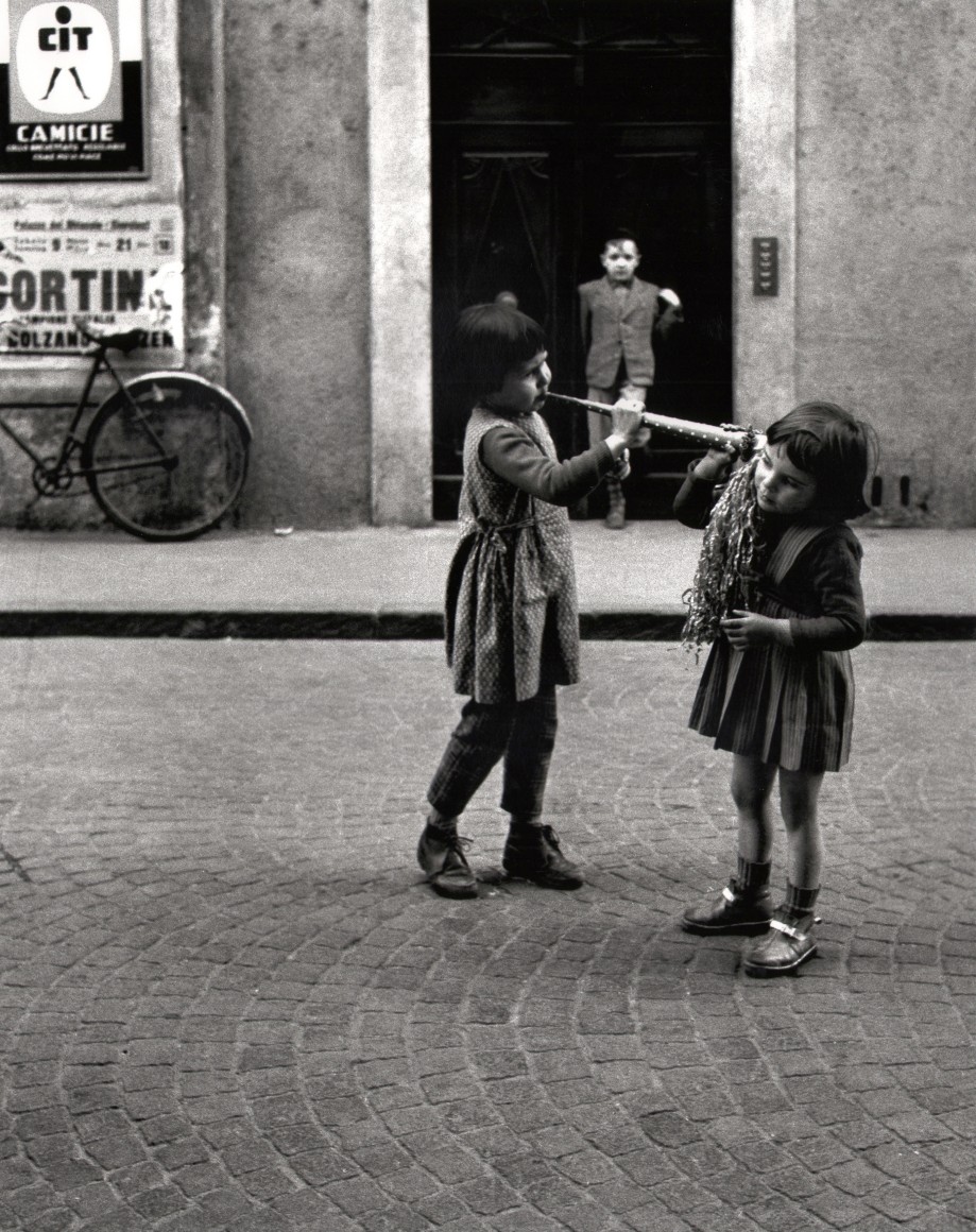 Nino Migliori, People of Emilia, 1950. Street scene featuring three children. One boy stands in a doorway in the background, while two girls play with a wind instrument in the foreground.