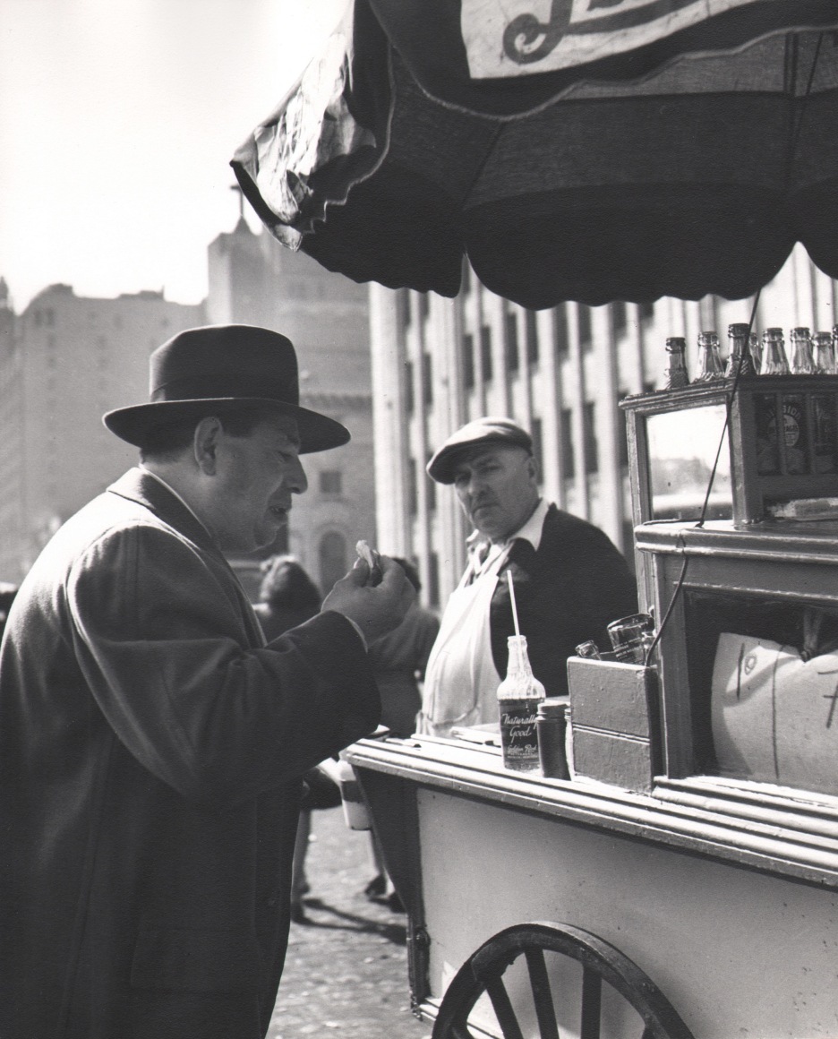 35. Esther Bubley, On South Street at noon time, 1946. Man in the left foreground in profile eating; man in the right midground stands in an apron behind a hot dog cart.