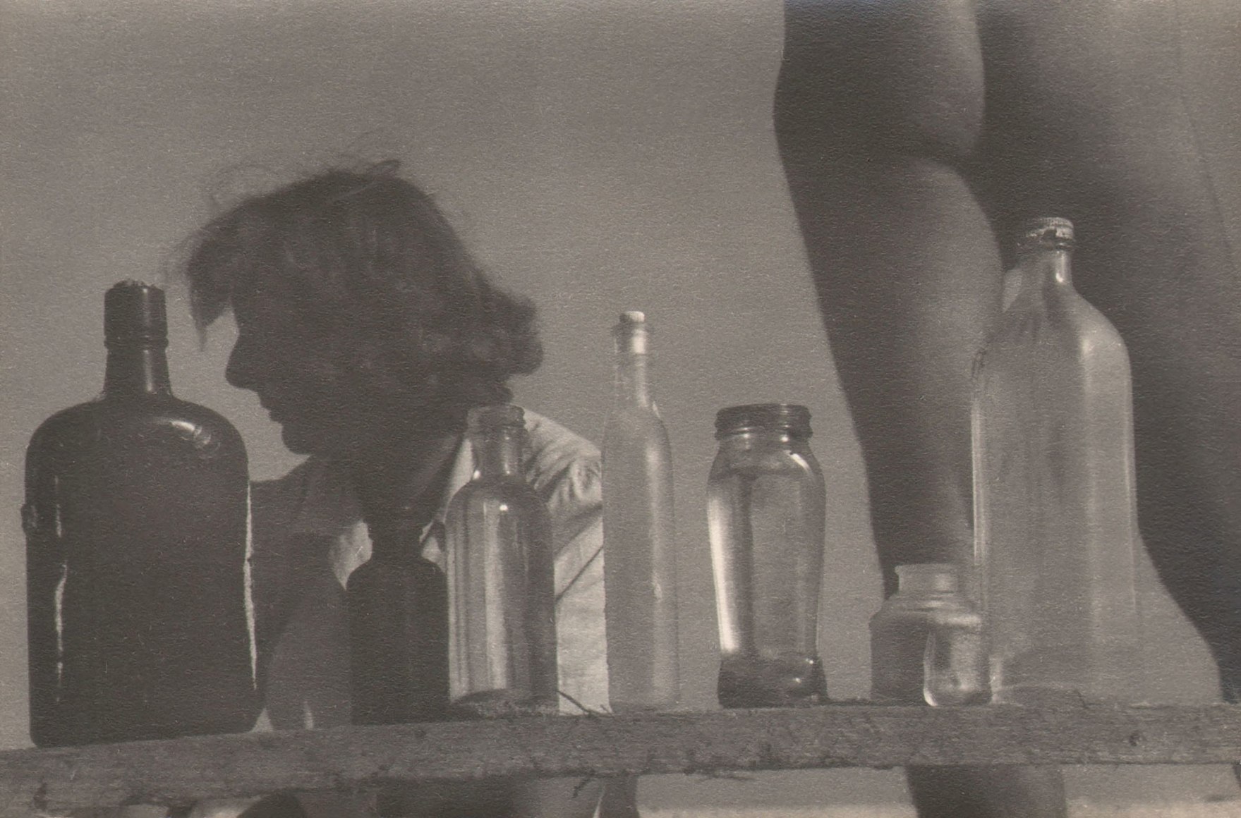 PaJaMa, Margaret French, Jared French, Fire Island, ​1945. A wooden plank extends across the lower frame with various glass bottles on it. The lower body of one nude figure, facing away, and the upper body of a woman facing left, are behind the bottles.