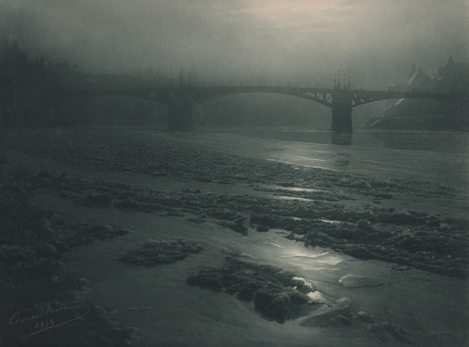 12. L&eacute;onard Misonne, Hiver &agrave; Dinant, 1934. Partially frozen body of water running under a bridge in hazy light. Gray/green-toned print.