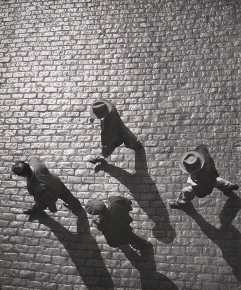 52. Bedrich Grunzweig, NYC, ​1948. Four men photographed from above, walking on a cobbled brick surface. Their shadows are cast down and to the right of the frame.
