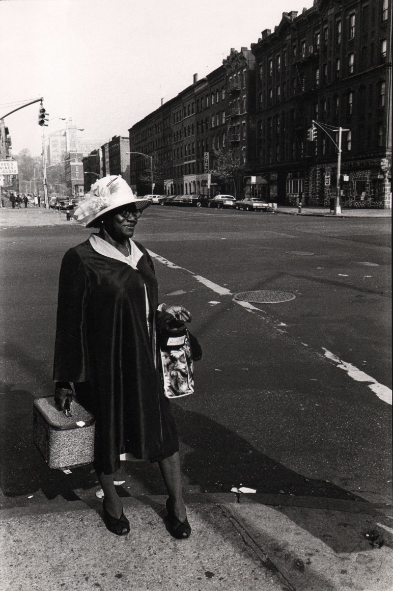 19. Anthony Barboza, Harlem, NY, ​1970s. A smiling, well-dressed black woman in a flowered hat smiles on the street corner.