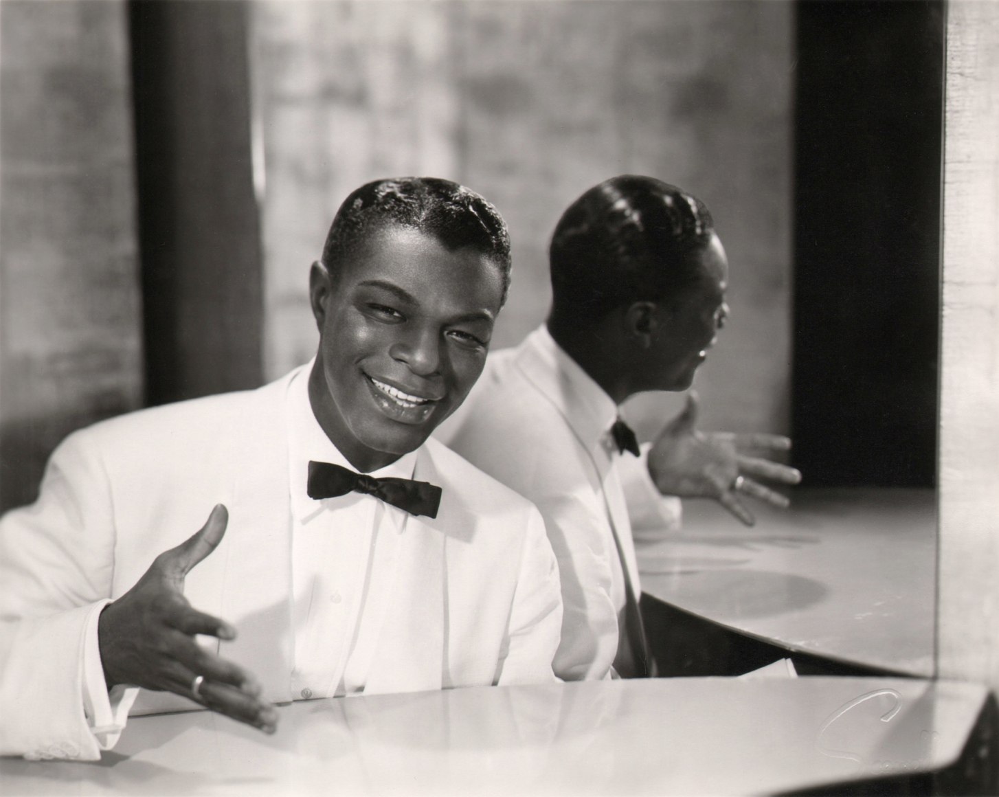 Wallace Seawell, Nat King Cole, ​c. 1955. Subject is seated in a white suit at the piano in front of a mirror, gesturing and smiling towards the camera.