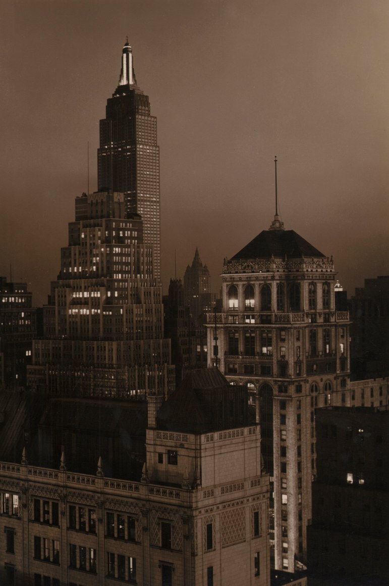32. Paul J. Woolf, Times Building Looking South, ​c. 1935. Night time sepia tone cityscape in a vertical composition. The Empire State Building is in the top left of the frame.